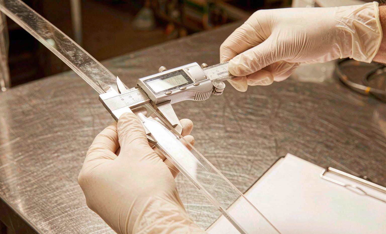 Gloved hands measuring the thickness of a PVC strip with a digital caliper on a steel workbench